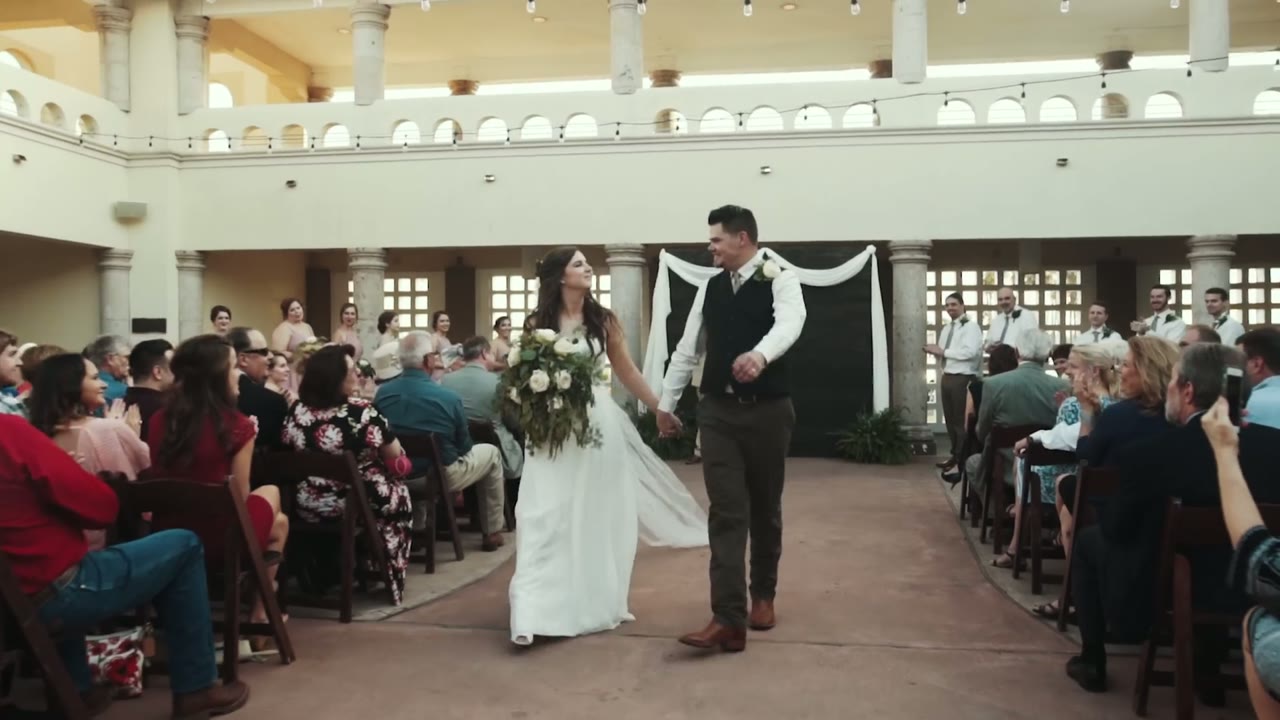 Bride and groom walking back up the aisle holding hands and smiling, guests seated on both sides in an arched courtyard
