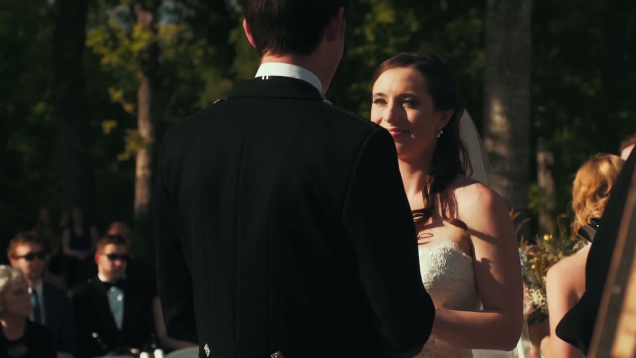 Bride smiling up at the groom in his kilted jacket during their vows, sunlight streaming through trees