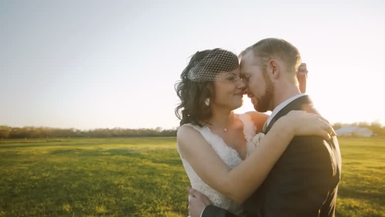 A gimbal shot of a bride and groom holding each other in a green field with the sun low behind them