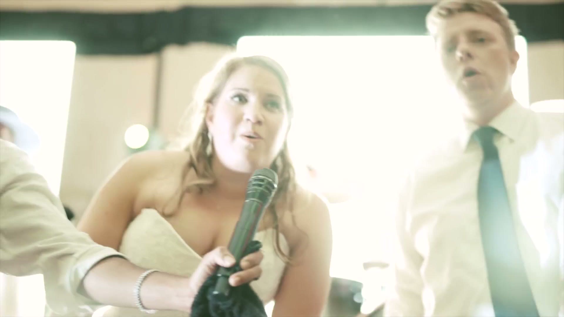 A bride holding a microphone and singing on a packed reception dance floor with the groom beside her