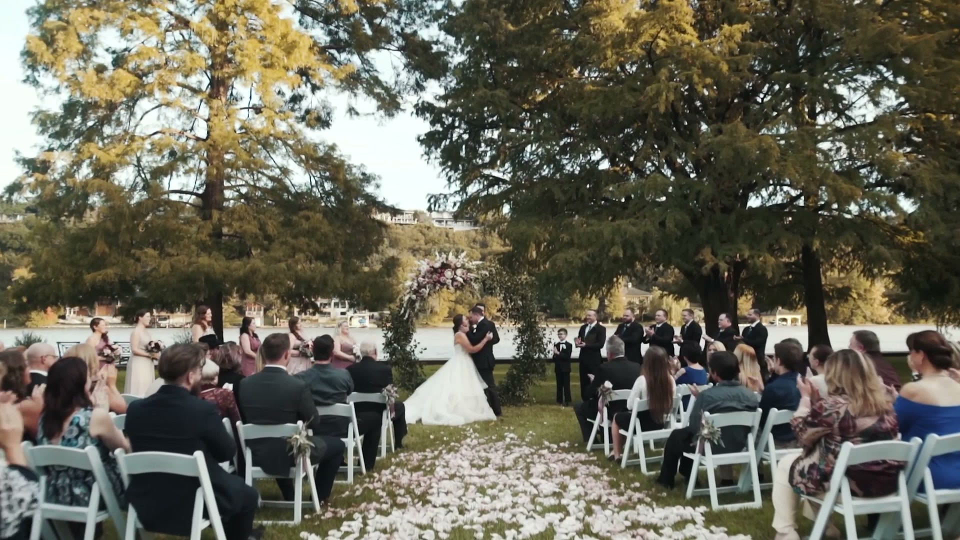 Wide shot of an outdoor wedding ceremony under tall trees beside a lake, guests seated along a petal strewn aisle