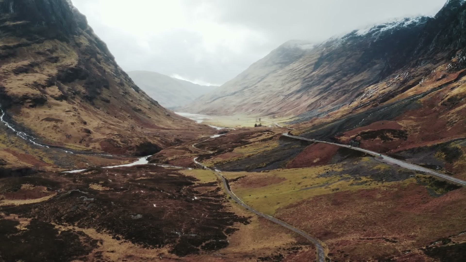 Aerial drone view looking down the Glencoe valley, with a river winding through red and gold moorland between two mountain ranges and a single road running along the right side