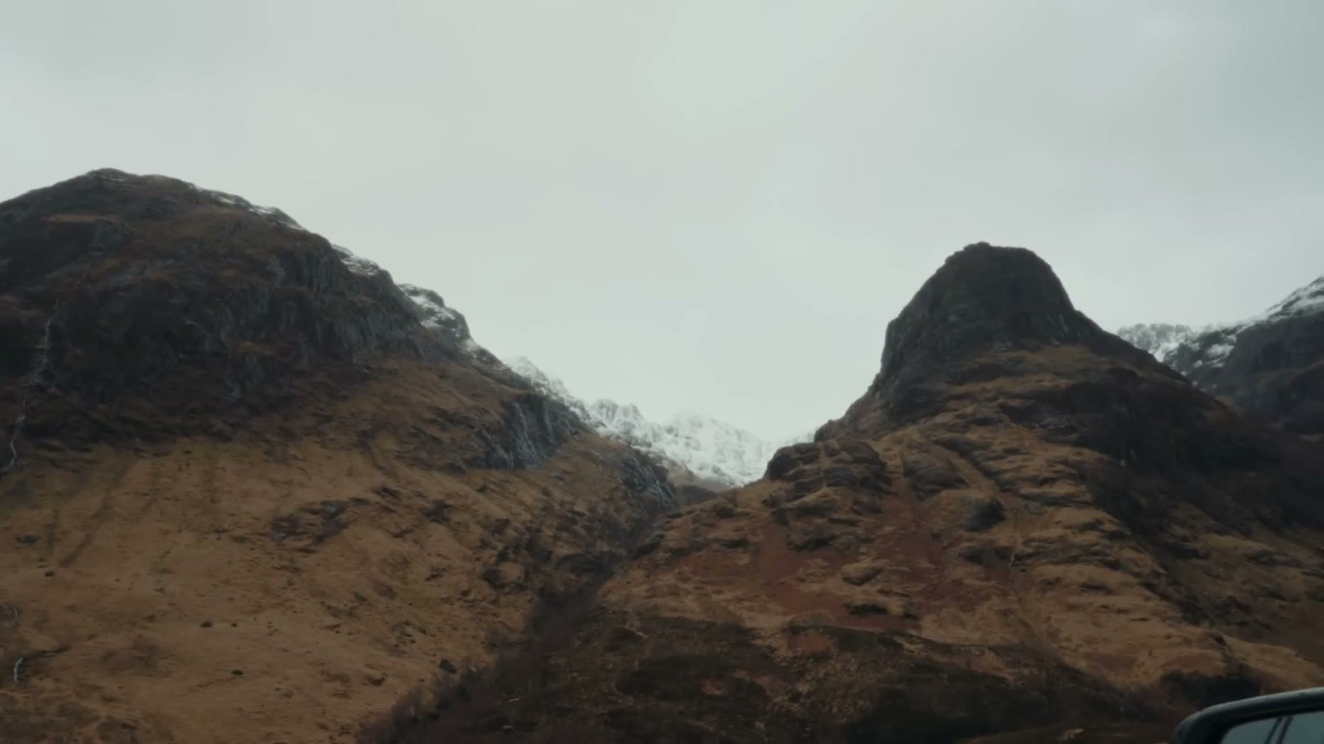 Two steep brown mountain peaks with snow on their upper ridges rising above the road at the Three Sisters viewpoint in Glencoe, Scotland