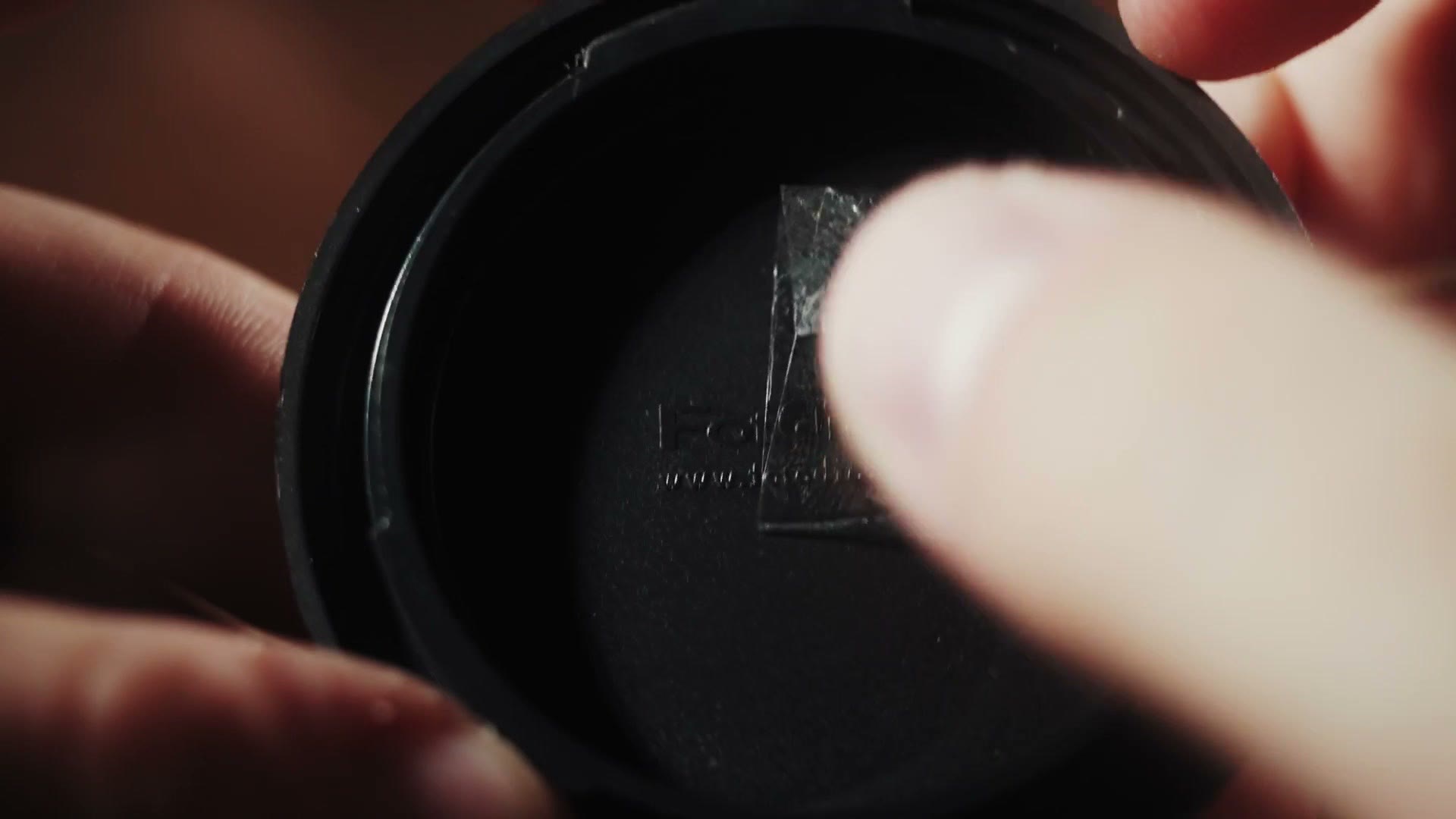 A thumb pressing a ring of clear double-sided tape firmly into the inside of a black camera body cap.