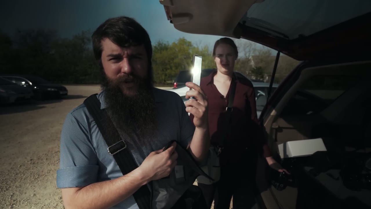 Close-up of Matt holding up a light diffuser prism at the car trunk, assistant with a shoulder bag behind him