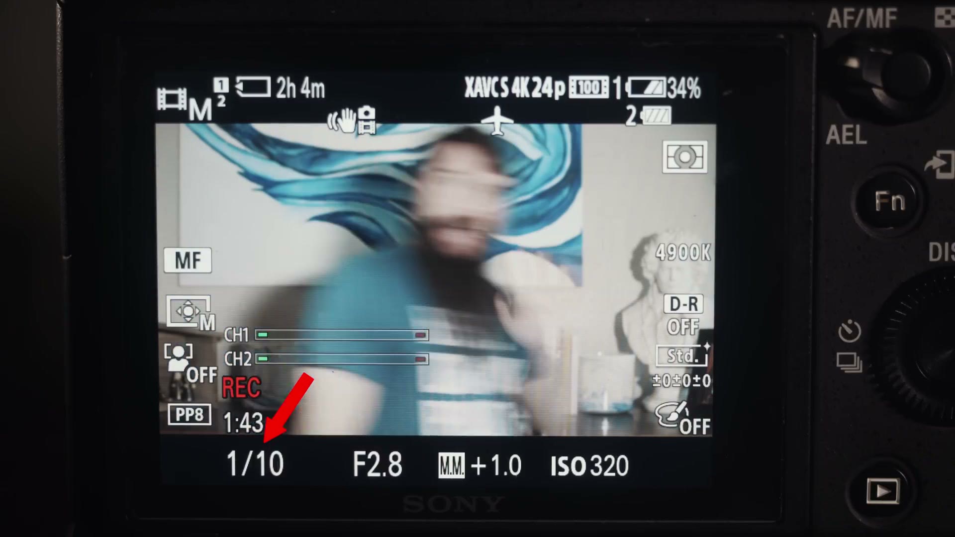 A camera rear screen with the shutter speed at 1/10 highlighted by a red arrow, and the live image behind it smeared beyond recognition