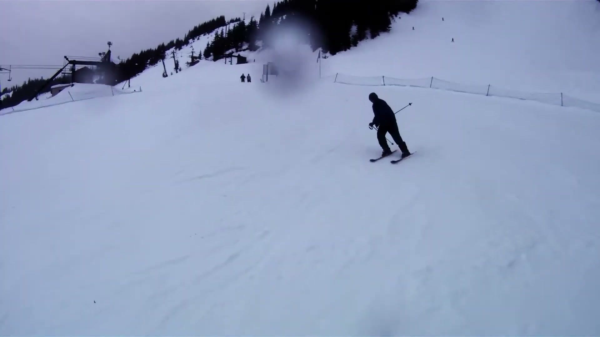 A skier in dark clothing traversing a wide snow slope under flat grey cloud, seen from a helmet mounted camera with water droplets visible on the lens
