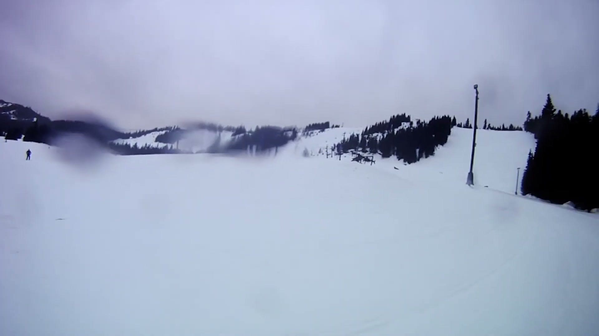 A wide view from a helmet camera across a snowy ski run toward a line of evergreen trees and a lift tower, with low cloud sitting on the ridge