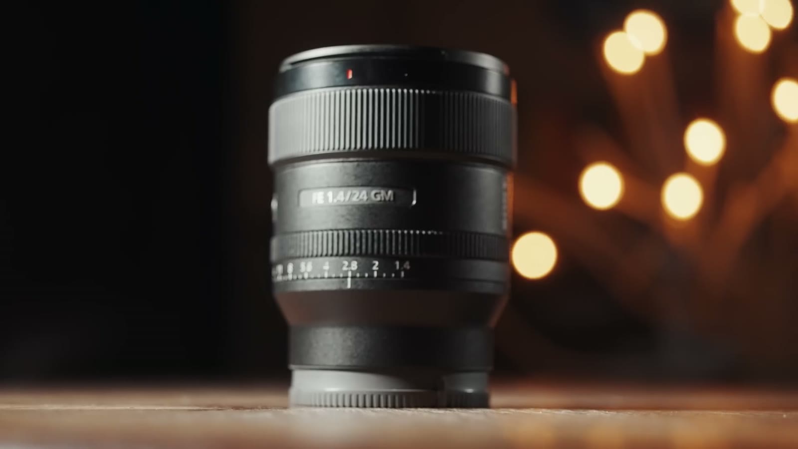 The Sony FE 24mm f1.4 GM lens standing on a table, its barrel marked FE 1.4/24 GM, with warm out of focus lights behind it