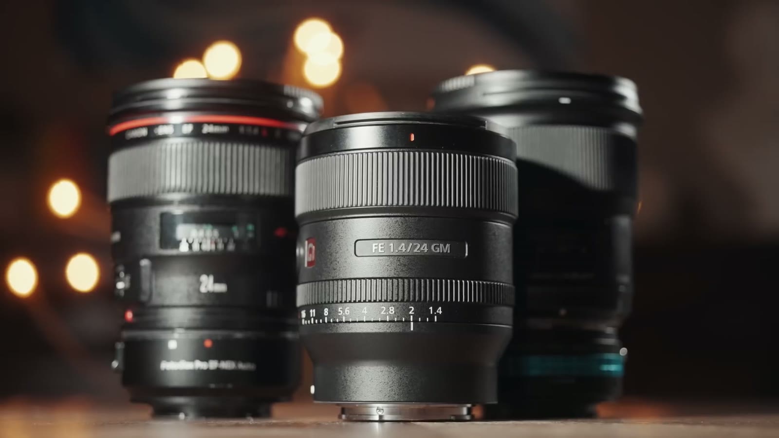 Three 24mm lenses side by side on a table, a Canon EF 24mm, the much smaller Sony FE 24mm GM in the middle, and a Sigma