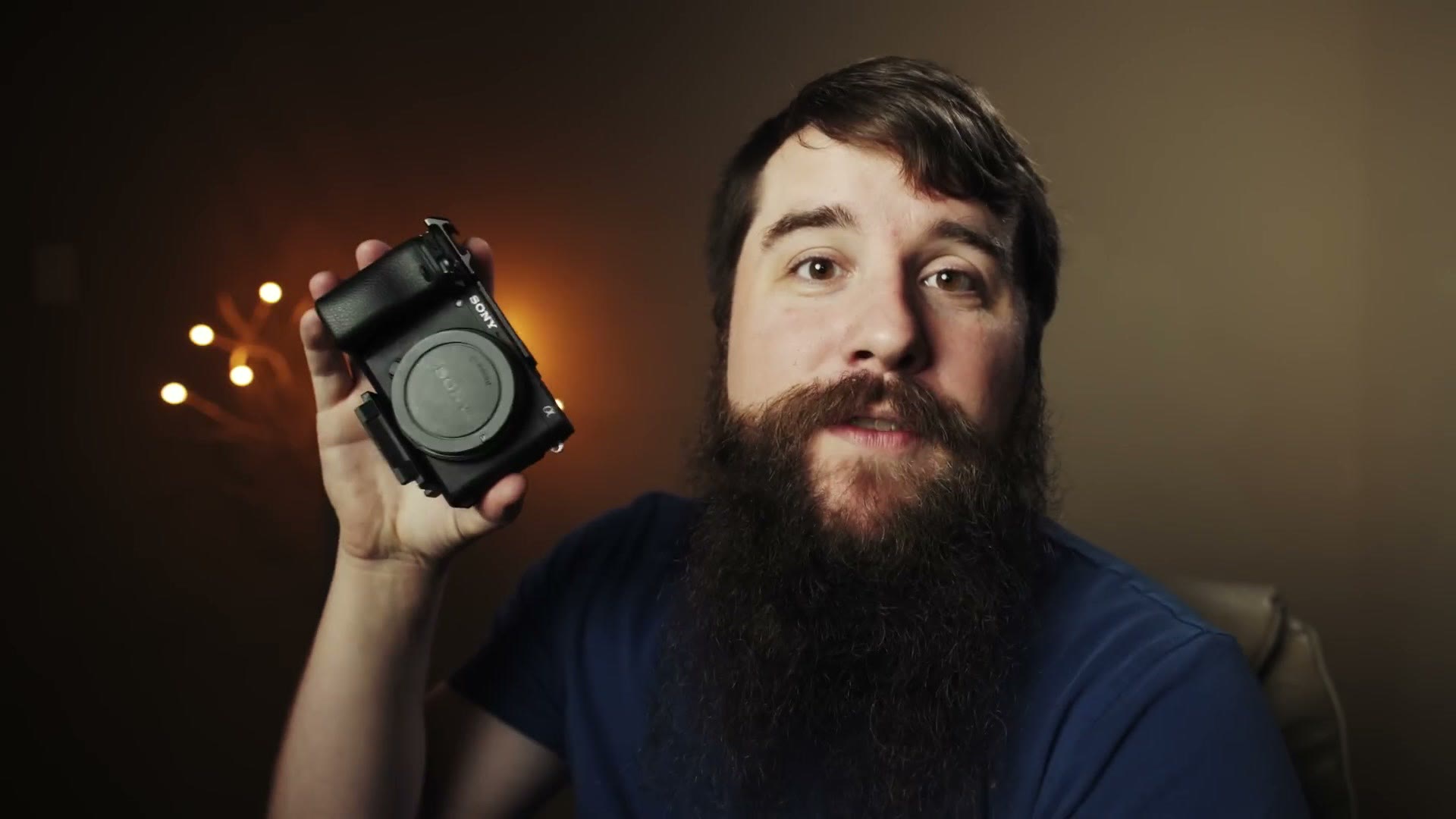 Matt holding a Sony a6400 up to the camera while talking, string lights glowing behind him