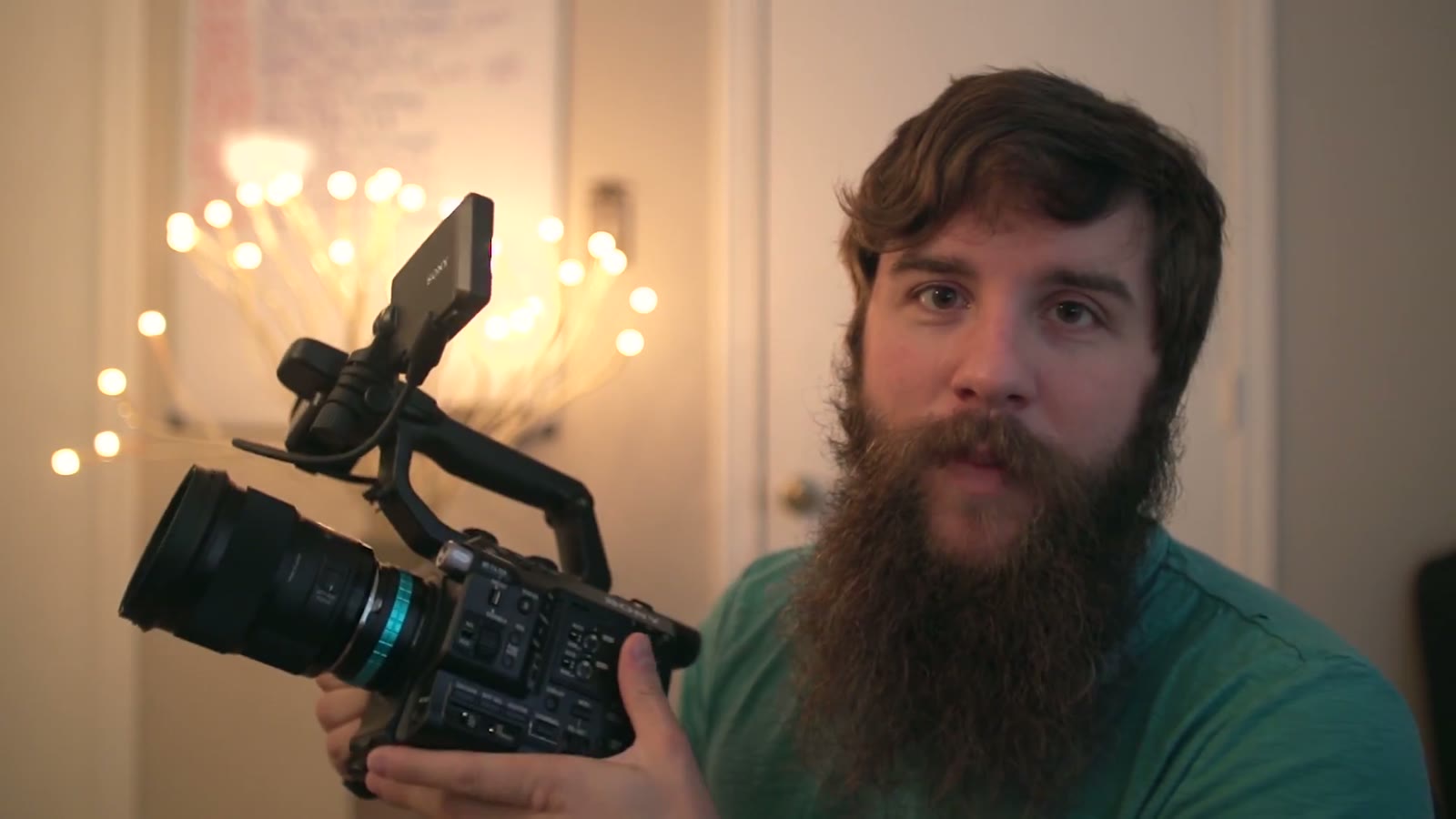 Matt holding the Sony FS5 close to camera with lens filling the foreground, string lights arcing behind him, mid-sentence