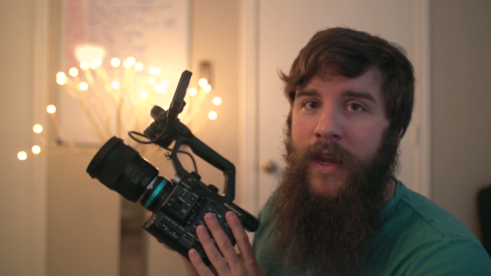 Matt holding the FS5 lower with more of its grip and side buttons visible, looking toward camera, string lights behind