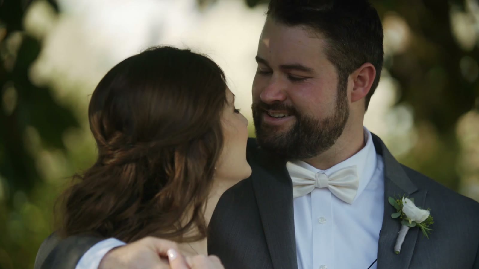 Bride and groom smiling close together outdoors under trees, groom in a cream bow tie with a white boutonniere