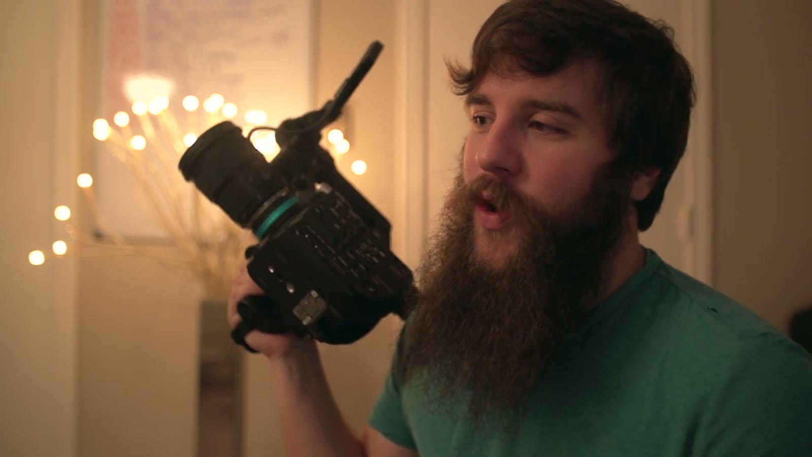 Matt speaking with mouth open mid-word, FS5 held up beside his face, string lights and a wall poster blurred behind