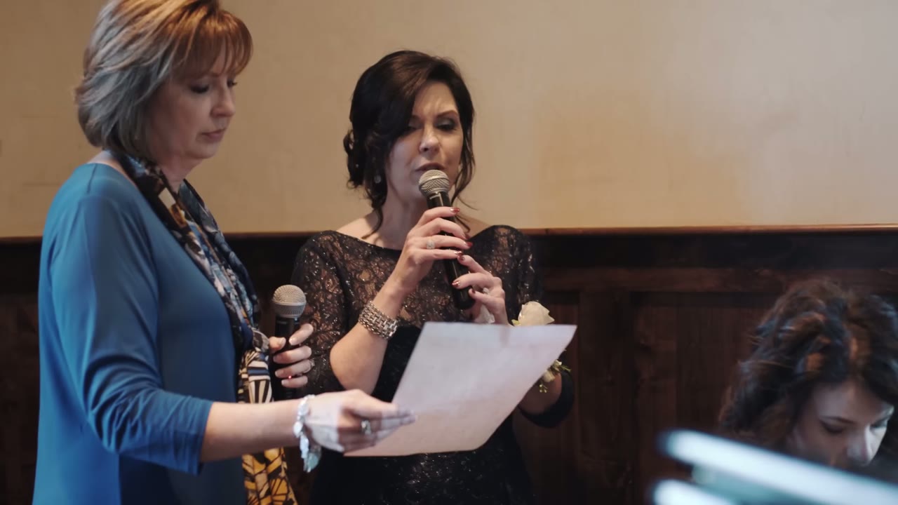 Two women at the reception sharing a microphone while one reads a speech from paper