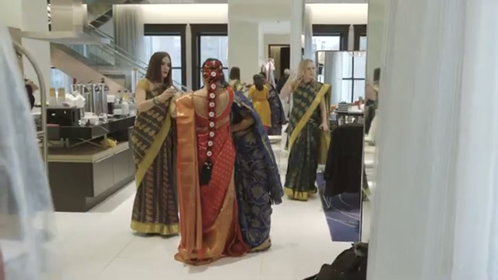 Bride in an orange and gold sari being dressed by bridesmaids in navy and green saris in a hotel lobby