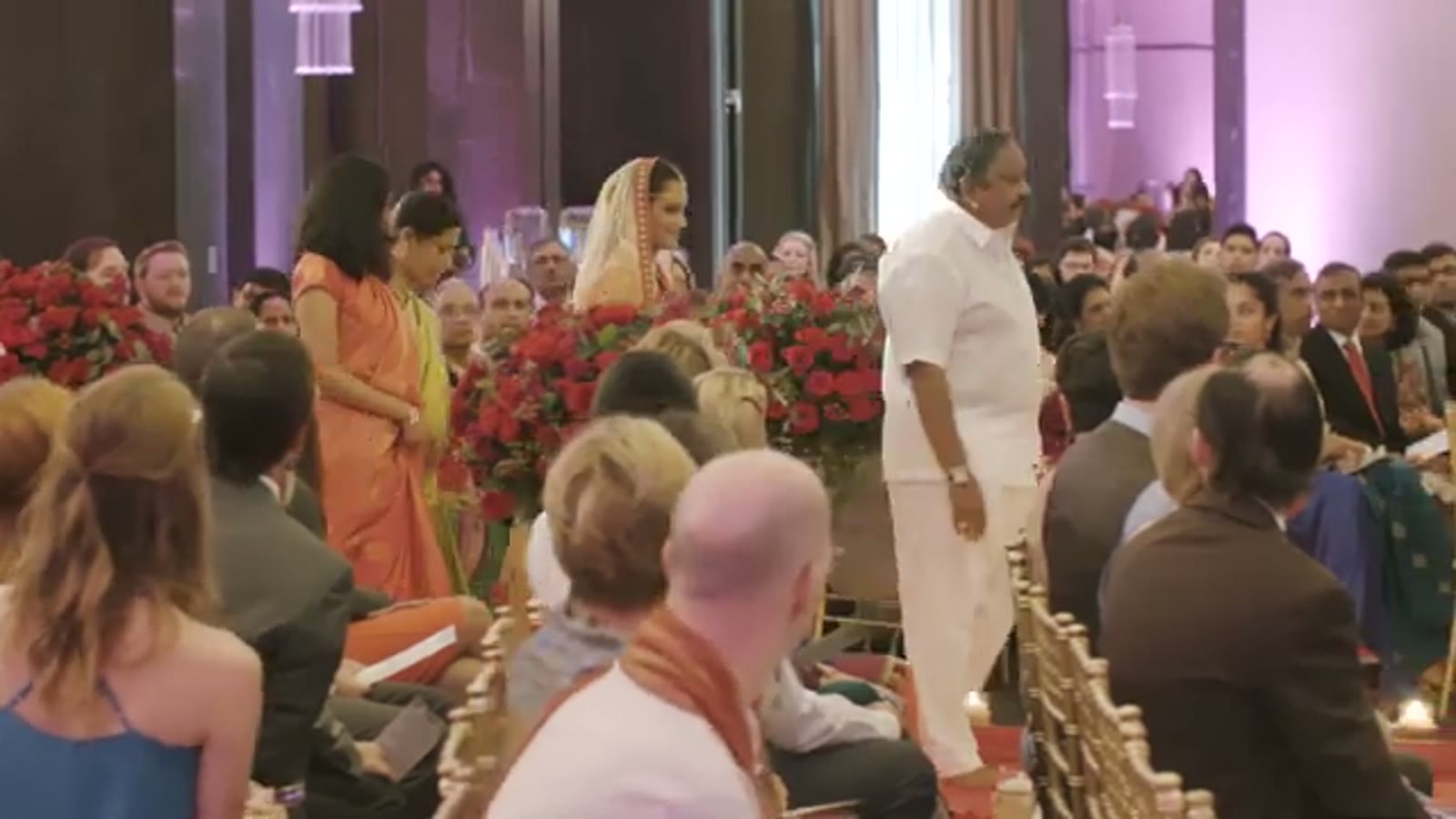Bride in a red veil walking down the aisle past red rose arrangements as seated guests turn to watch