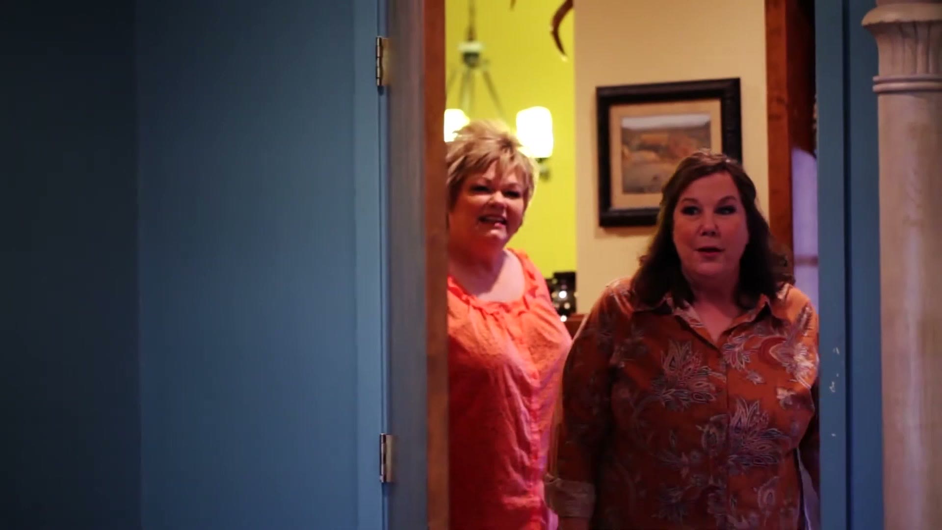 Two women standing in an open doorway, both mid-reaction with wide eyes, having just walked into a room they were told held a photograph