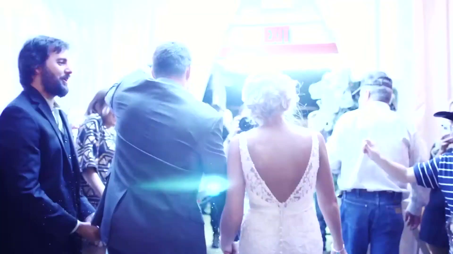 The bride and groom walking away from camera through a crowd of guests in a bright hall, guests reaching out on both sides