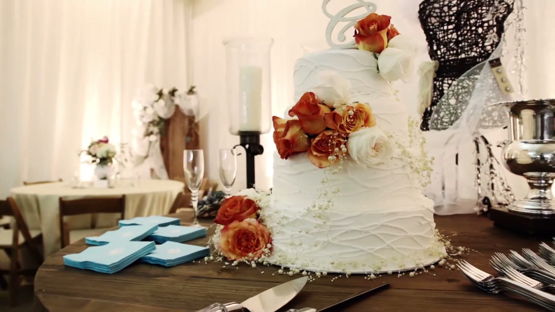 A white tiered wedding cake decorated with orange roses on a wooden table, with turquoise napkins, champagne flutes and a cake knife beside it