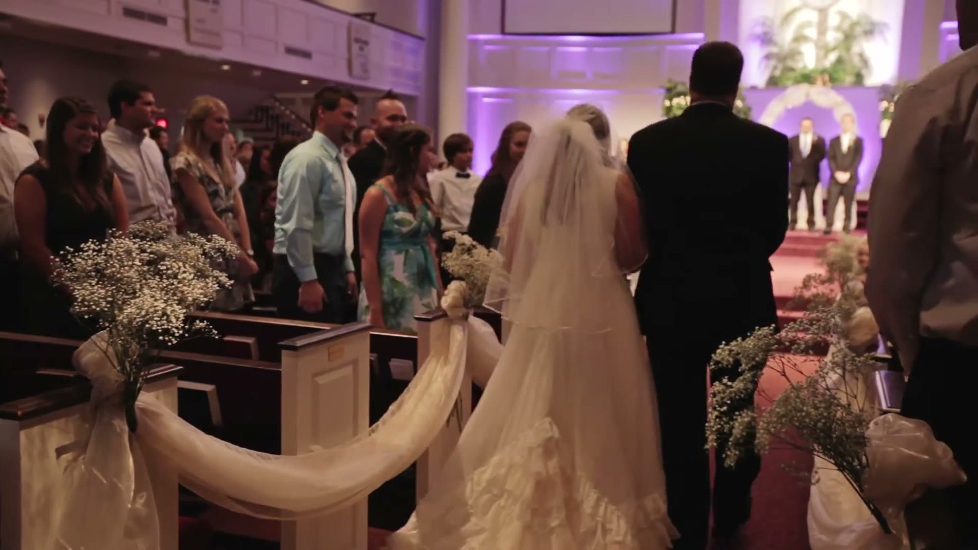 A bride in a veil walking down a church aisle on her father's arm, guests standing in the pews on either side, purple uplighting on the walls ahead