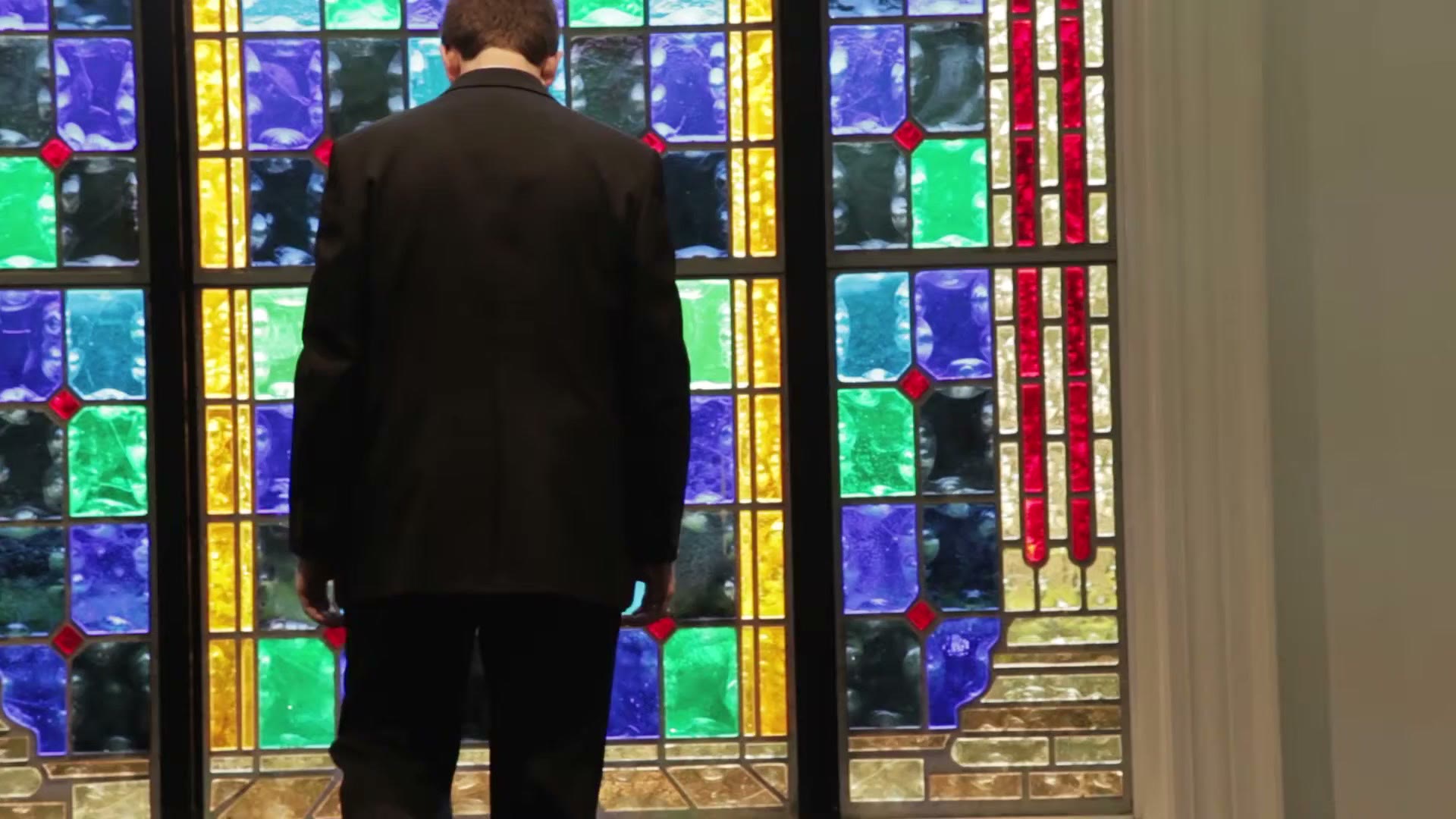 A groom in a dark suit standing with his back to camera in front of a tall stained glass window in shades of blue, green, yellow and red