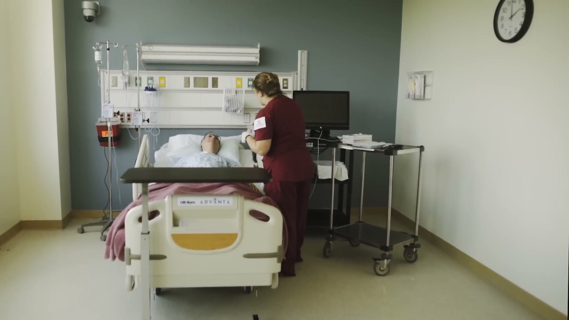 A student nurse working at a bedside in the simulation ward, with a high fidelity mannequin in a hospital bed and full monitoring equipment on the wall