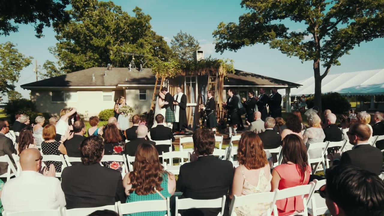 Wide shot of a backyard wedding ceremony under a wooden arch, guests in white folding chairs facing the couple