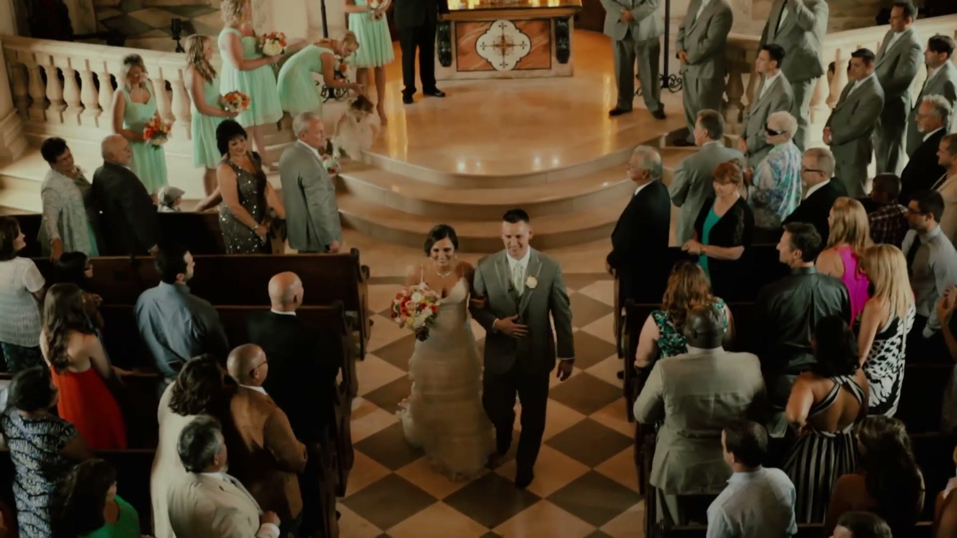 A bride and groom walking back down a church aisle arm in arm on a black and white checkerboard floor, guests standing and applauding on both sides