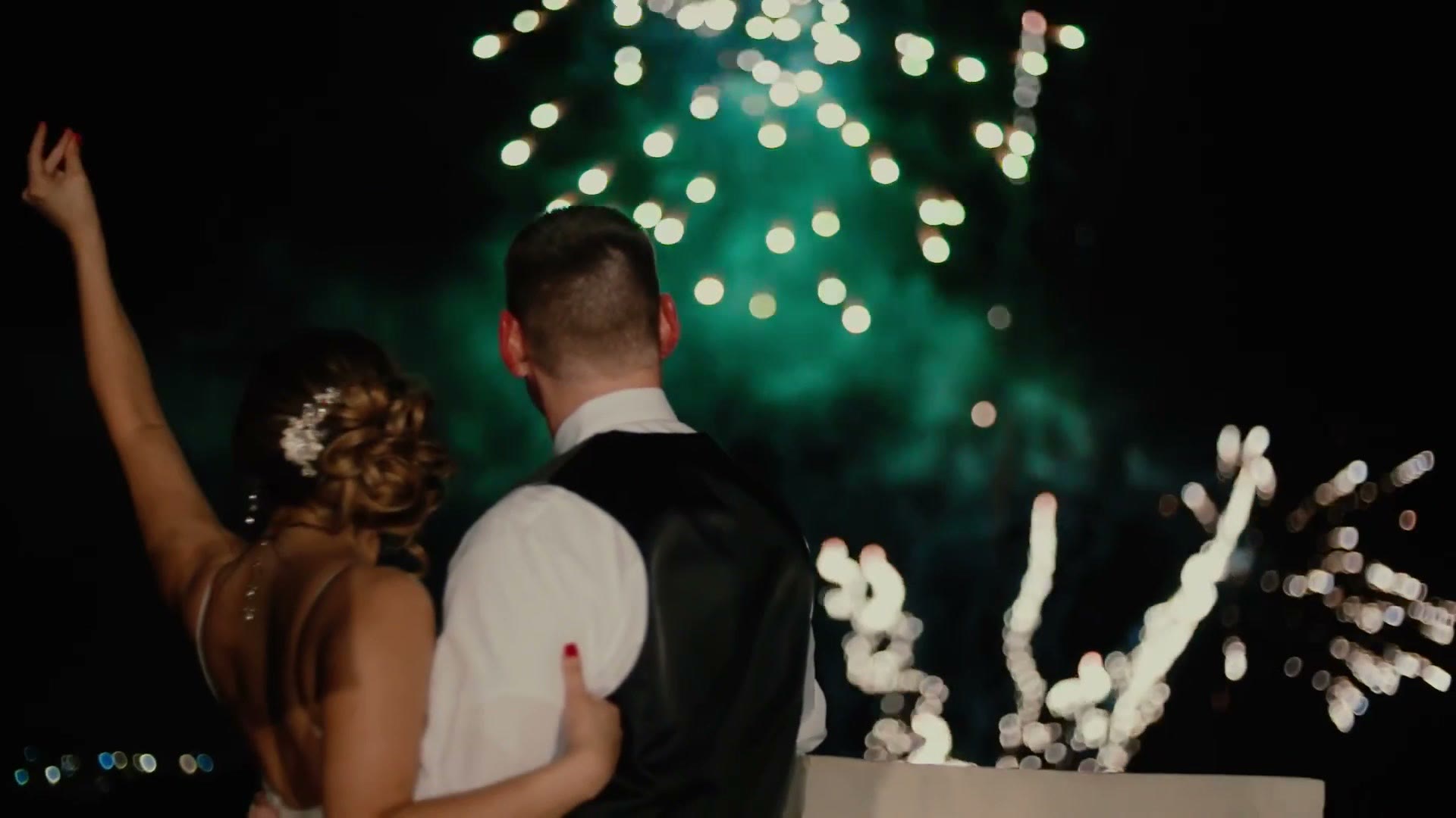 A couple dancing outdoors at night with fireworks bursting in the sky behind them, the bride's arm raised