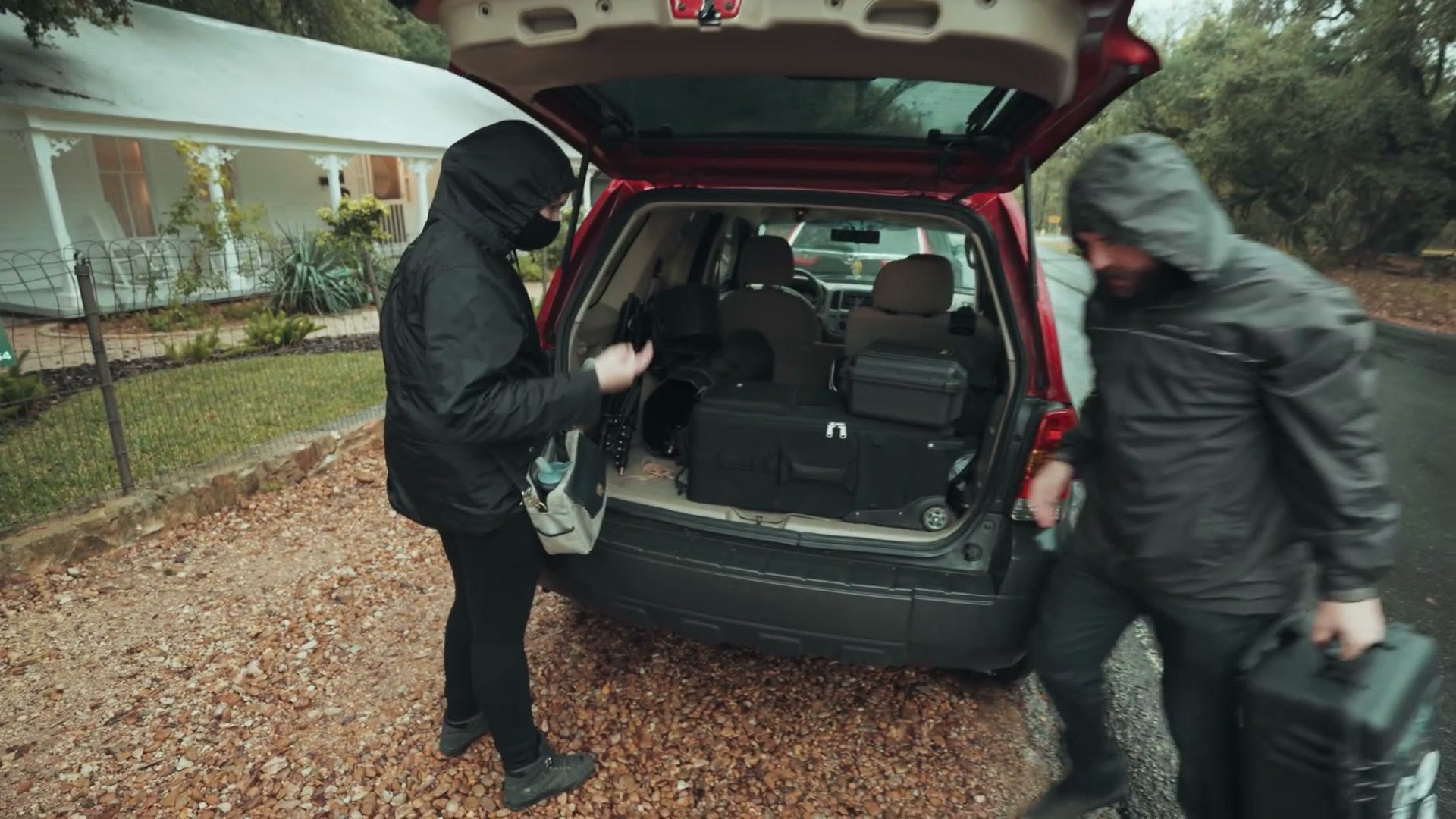 Two masked crew members unloading camera cases from the open trunk of a red SUV in the rain