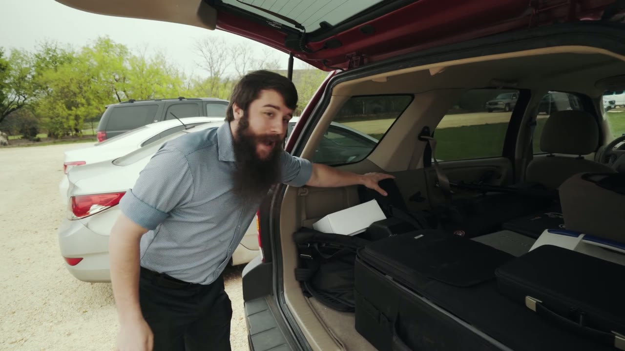 Matt leaning into an open car trunk packed with camera cases, talking to the camera about the gear