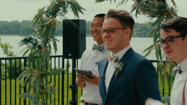 The groom in a blue suit and round glasses waiting at the front with his groomsmen under a greenery arch, the lake behind them