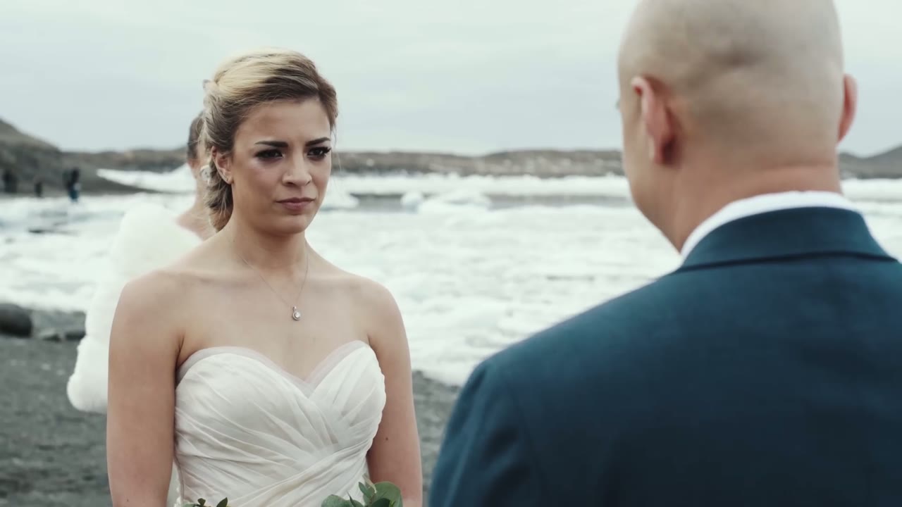 Bride and groom facing each other on a black-sand beach with icebergs floating in the water behind them