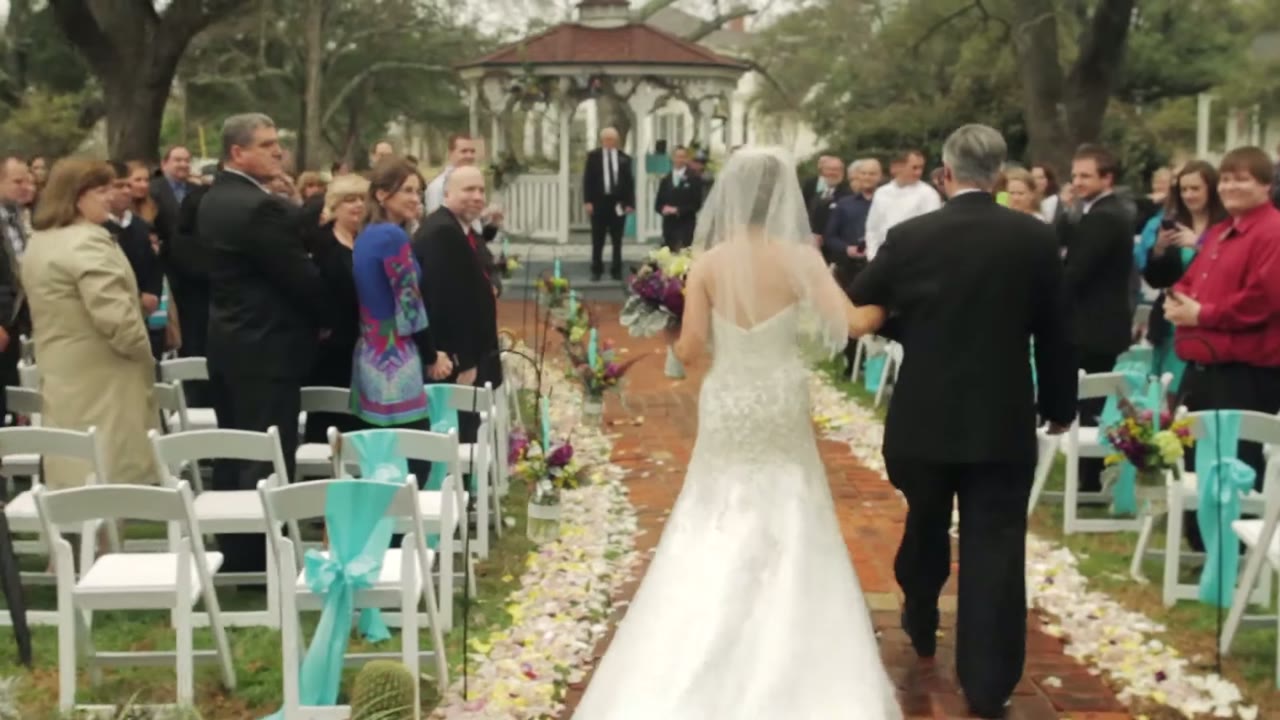 Bride walking down the outdoor aisle with her father, guests seated in chairs tied with teal ribbon