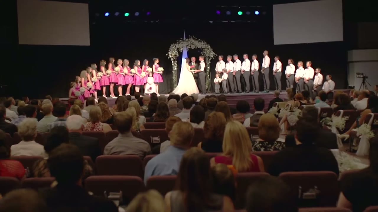 Wide view from the pews of the wedding party lined up on stage under colored stage lights