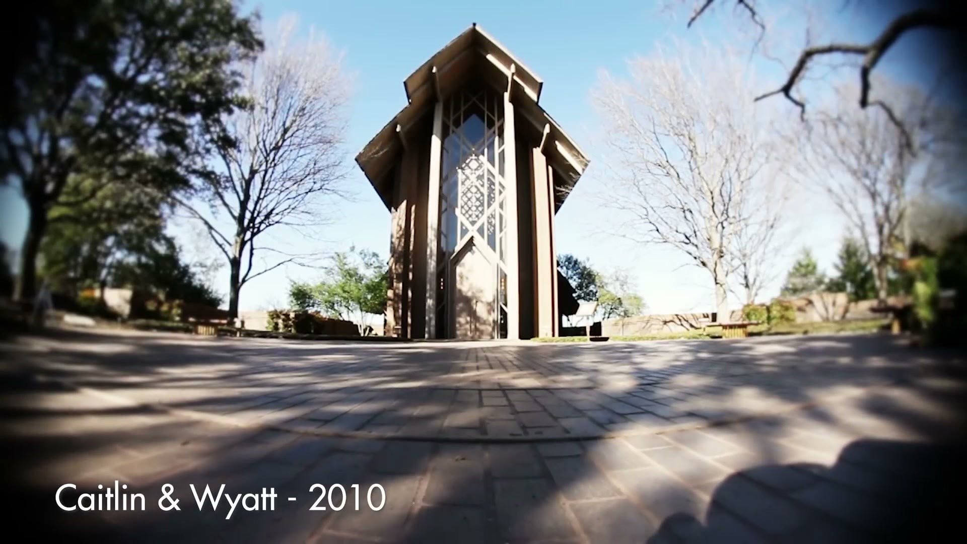 A wide fisheye shot of a tall wooden and glass chapel, with an on-screen caption naming the couple and the year 2010.