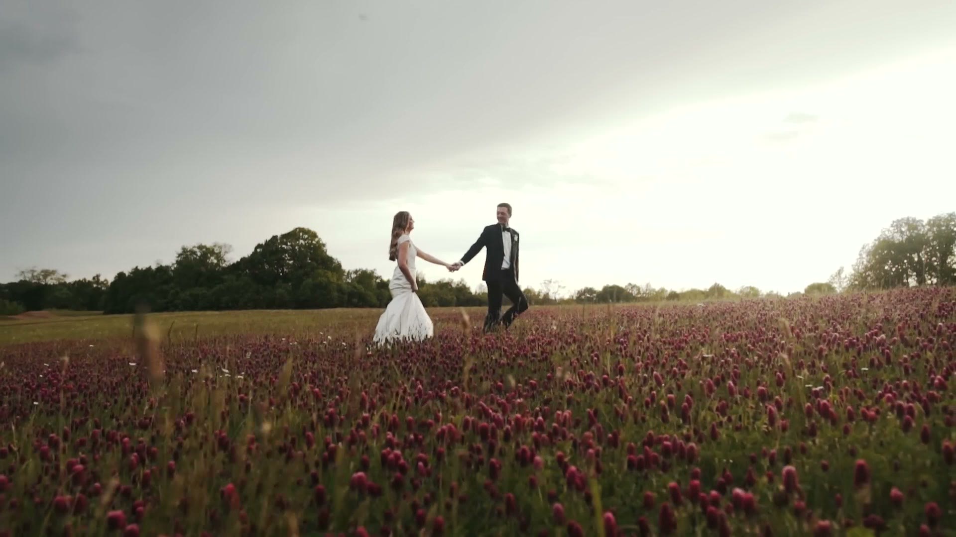 A bride and groom walking hand in hand through a field of crimson clover at golden hour.