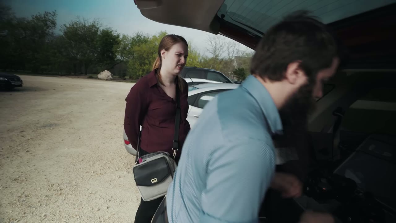 Wide shot of Matt and an assistant loading gear into an open car trunk in a gravel parking lot