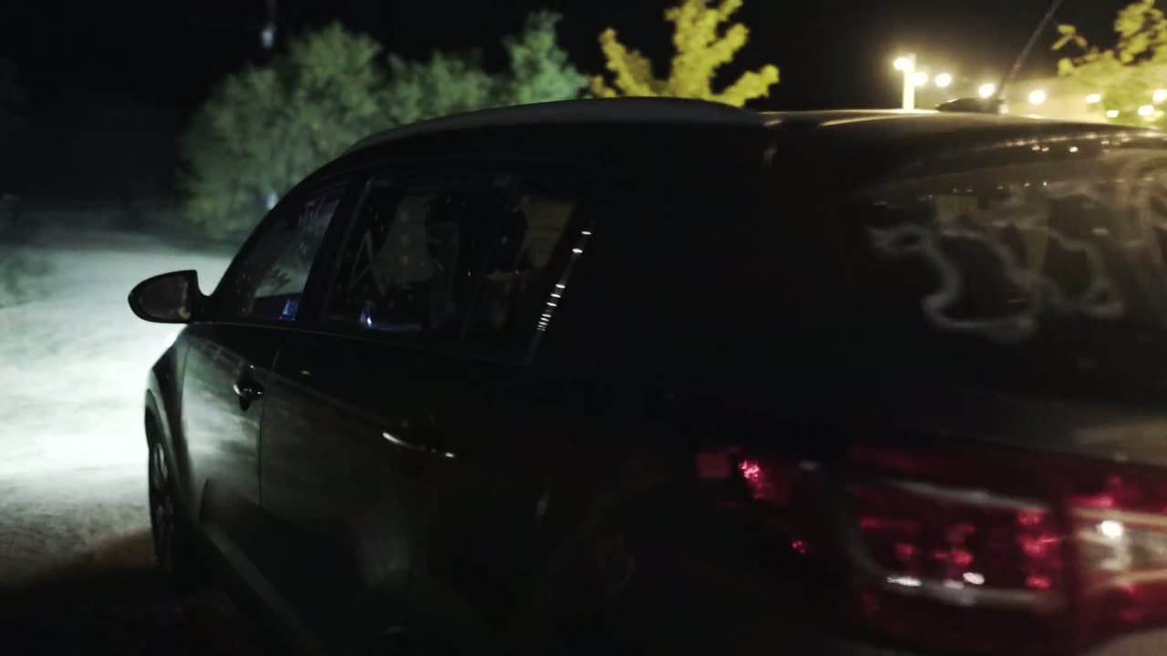 Dark getaway car at night with writing scrawled on the rear window, string lights glowing in the background