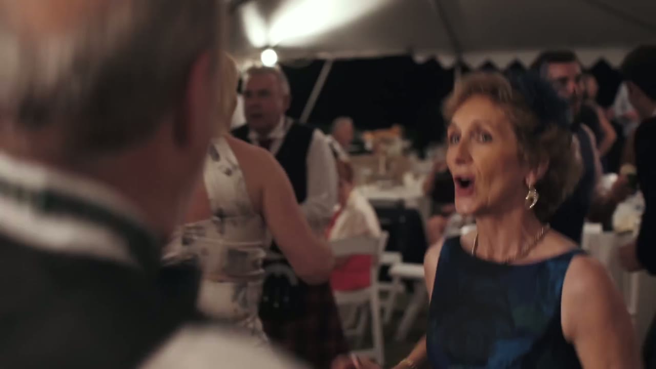 Wedding guest under the reception tent reacting with an open-mouthed laugh, a man in a kilt behind her