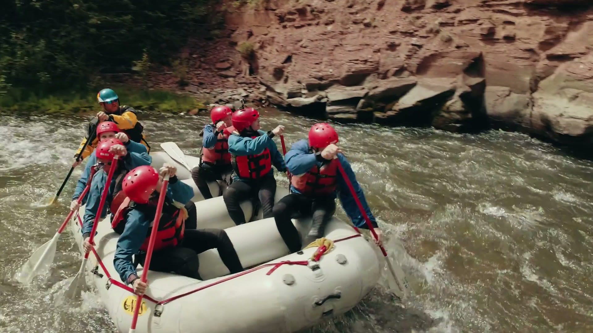 Rafters in red helmets paddling through rapids beside a red rock canyon wall on the San Miguel River