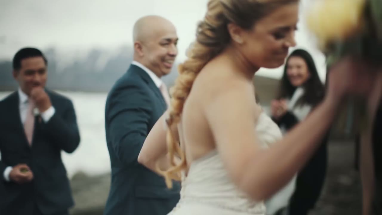 Bride and groom walking away from camera after the ceremony, guests smiling and clapping on a stony shore