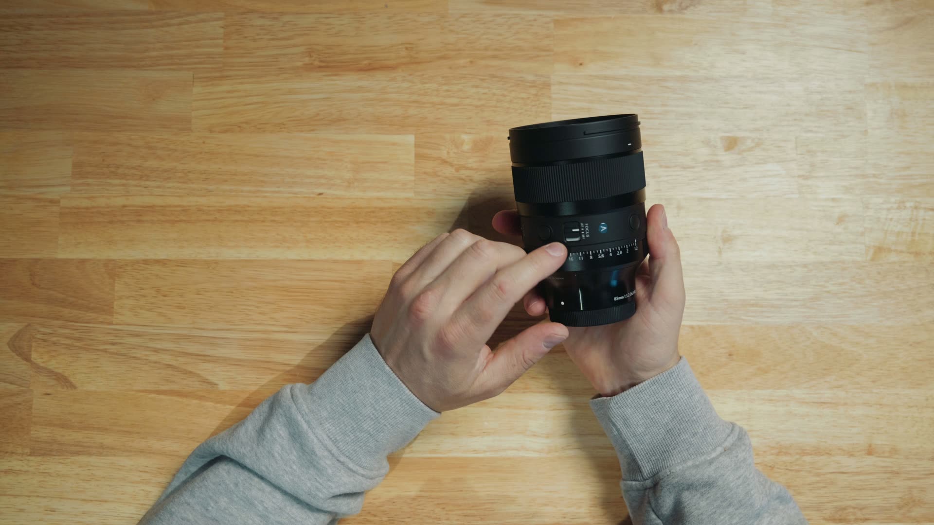 Overhead view of the Sigma 85mm F1.2 DG Art lens held on a wooden desk, showing the focus mode switch, custom buttons and aperture ring